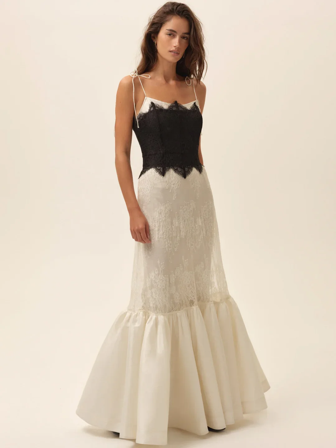 A woman wearing a long, elegant gown with a black lace top and a cream-colored, ruffled bottom, standing against a plain background.