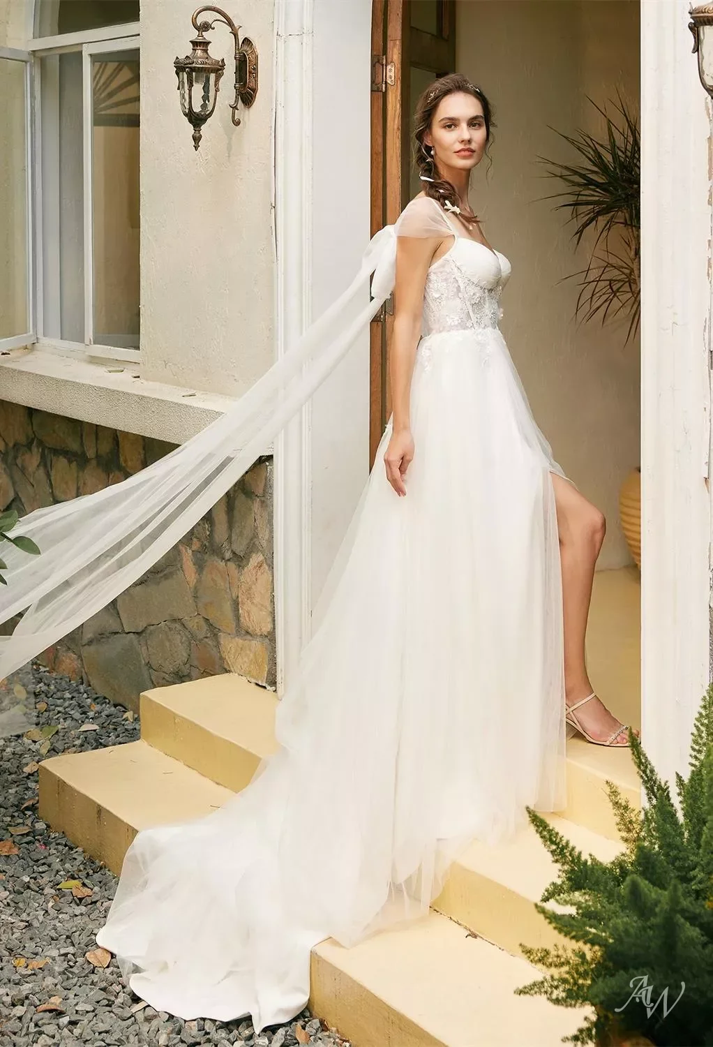 A woman wearing a wedding dress standing at the top of a small staircase outside a house, with a flowing veil, floral earrings, and strappy heels.