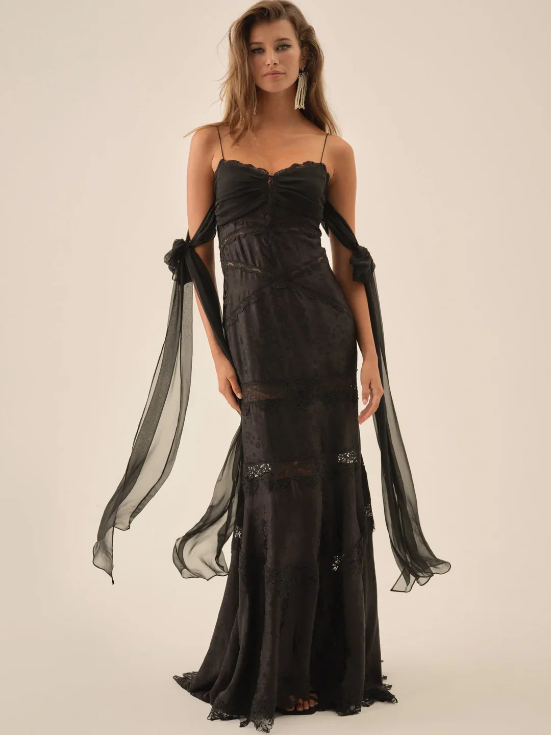 A woman wearing a long, black lace dress with spaghetti straps and sheer, flowing sleeves tied into bows.