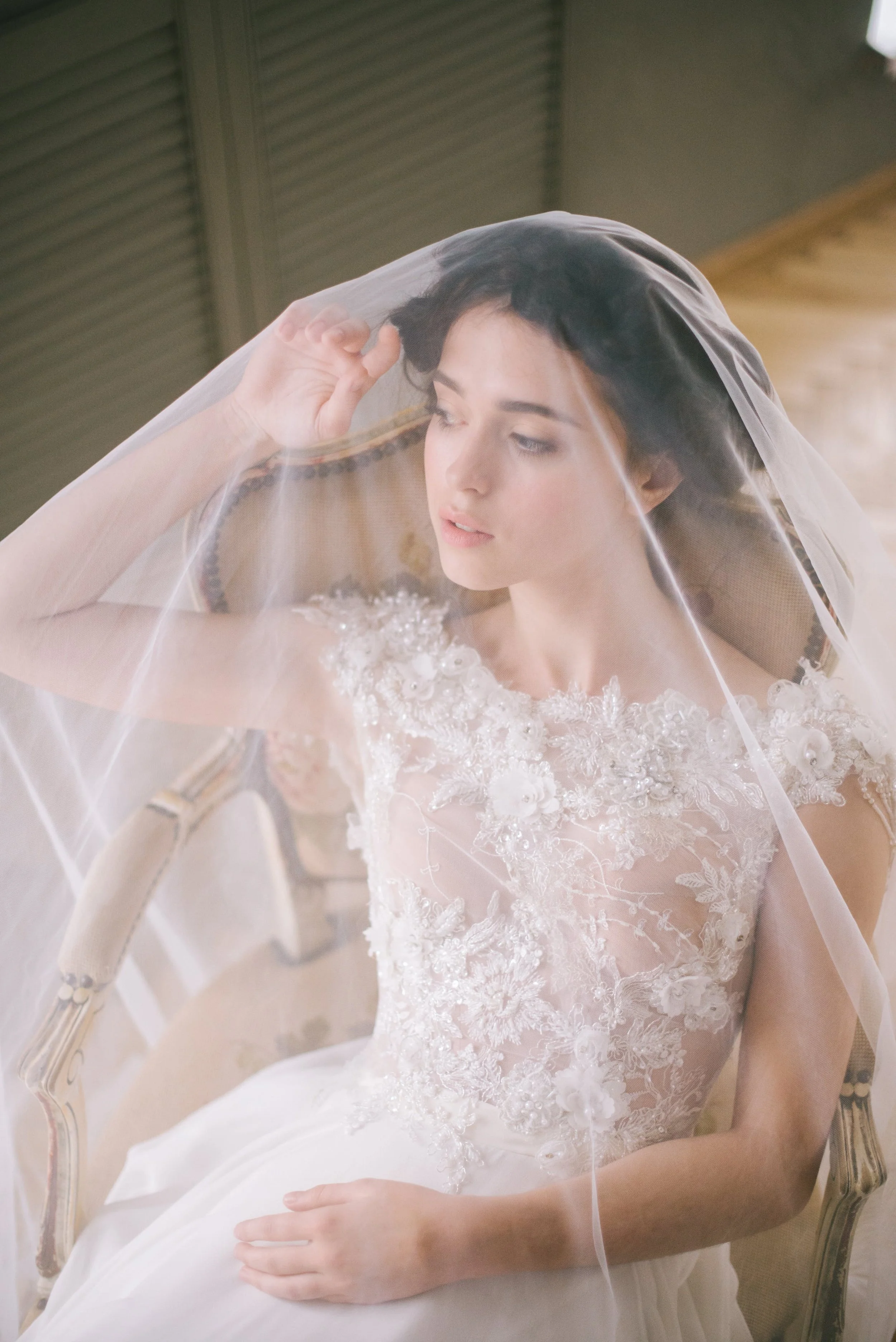 A bride with dark hair wearing a lace wedding dress sitting on a vintage armchair with a sheer veil draped over her head, indoors with wooden flooring and paneling in the background.