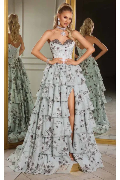 A woman in an elegant, strapless, floral gown with layered ruffles and a thigh-high slit, standing in front of a mirror, wearing jewelry and striking a pose.