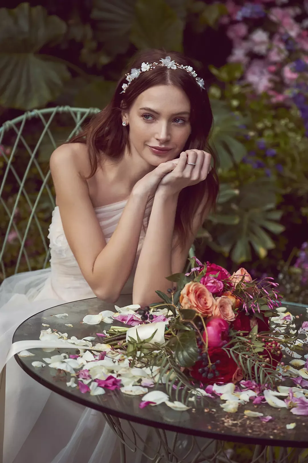 A woman in a white dress, wearing a floral headband and earrings, is sitting at an outdoor table decorated with flower petals and a bouquet of pink and orange flowers, surrounded by lush greenery.