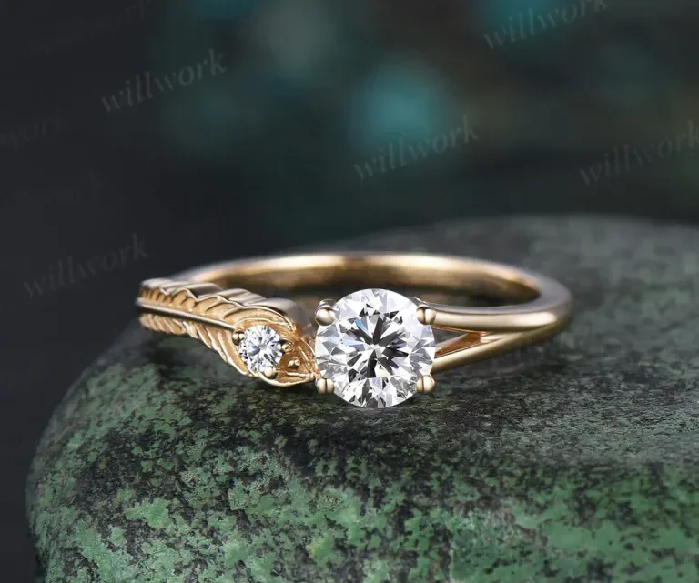 Gold ring with a large central diamond and a smaller diamond on an engraved leaf-shaped band, placed on a textured green stone surface.