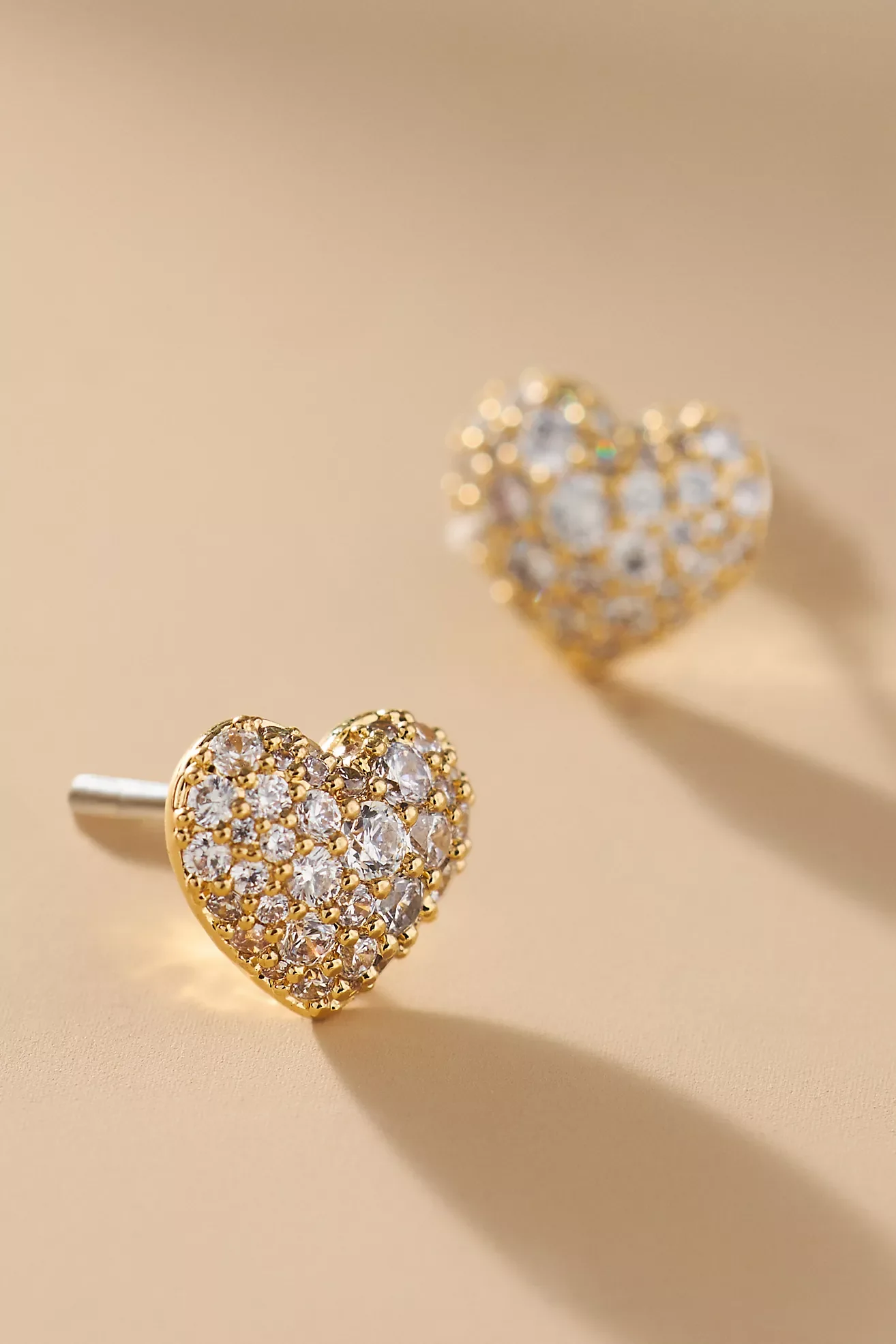 Gold heart-shaped stud earrings encrusted with small diamonds placed on a beige background.