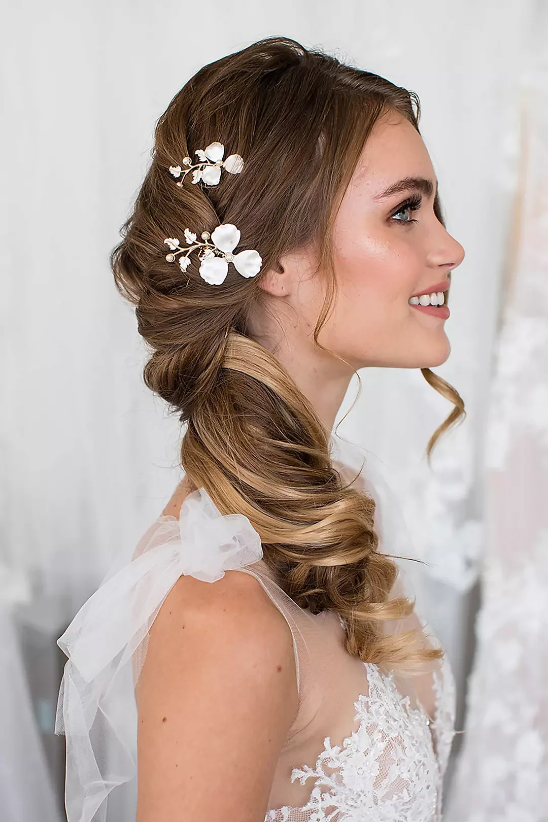 A young woman with styled hair adorned with white floral hair accessories. She is smiling and wearing a white lace dress.