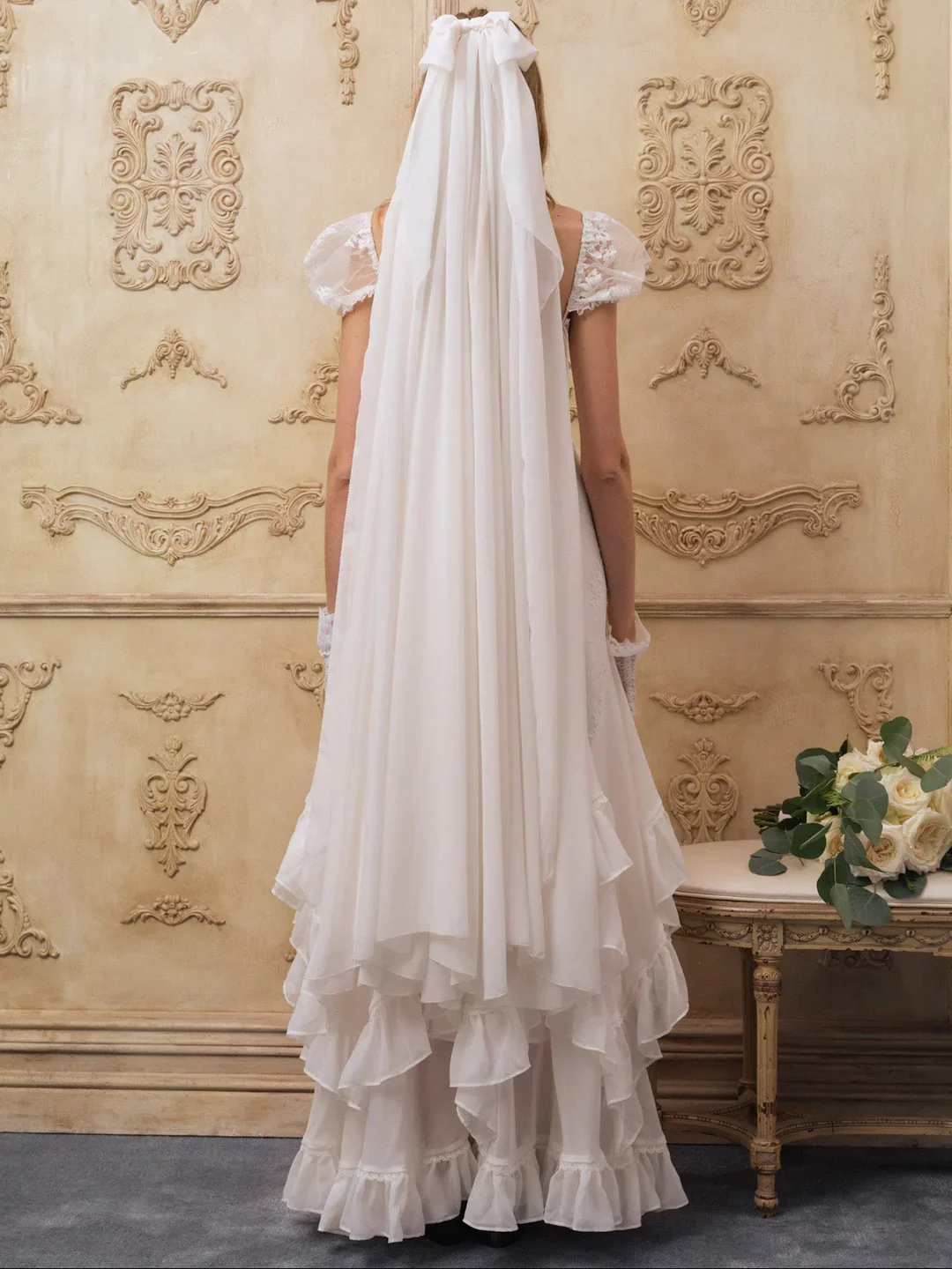 A woman wearing vintage white wedding dress with lace puff sleeves, standing in a decorated room with ornate wall designs, holding a bouquet of white roses and eucalyptus.