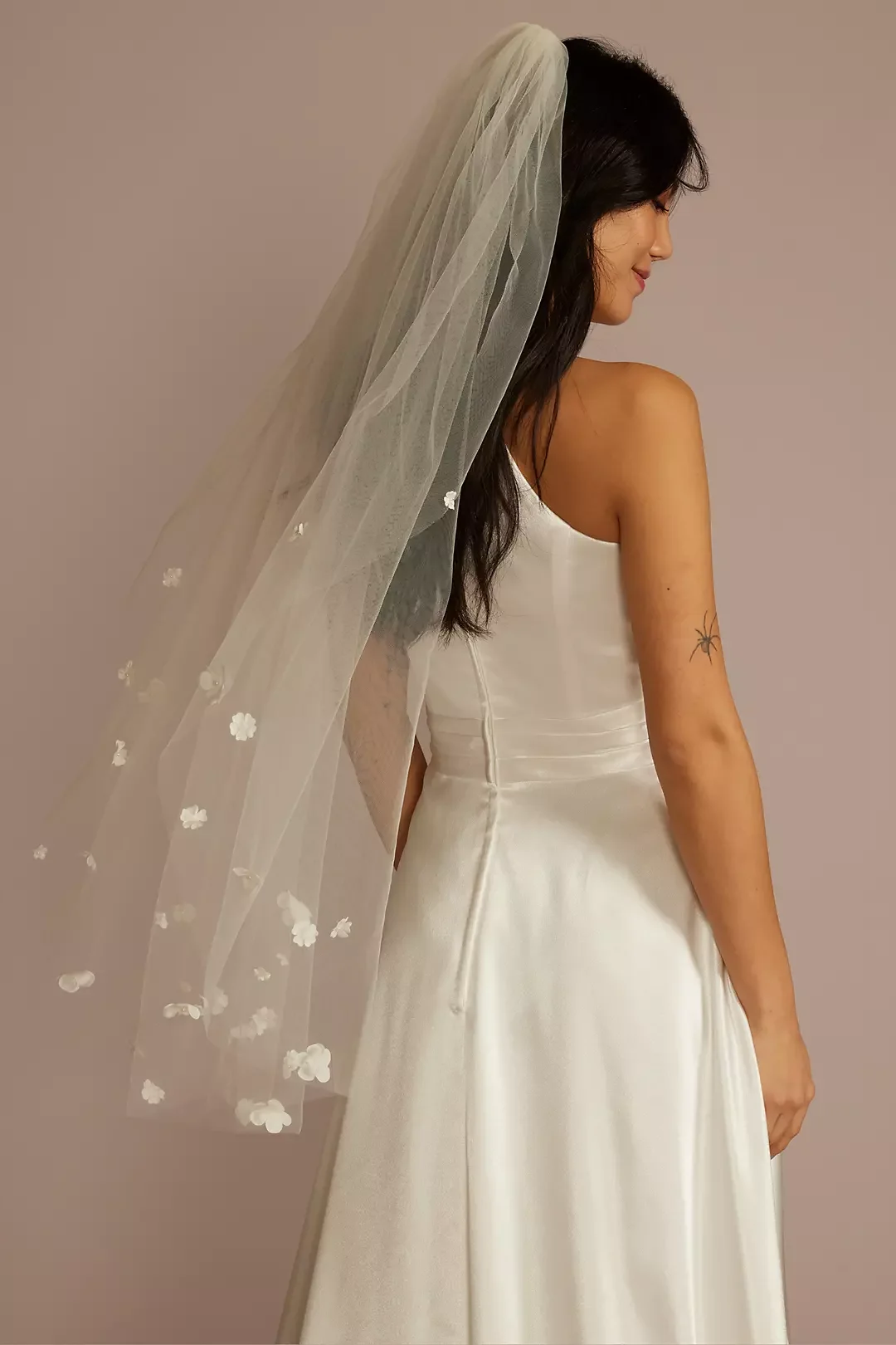 Woman in a white wedding dress with a veil, showing her profile and tattoo of a spider on her arm, against a plain background.