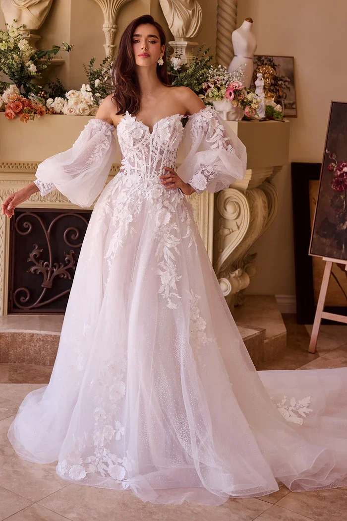 A woman in an ornate wedding dress with off-the-shoulder puff sleeves, lace details, and a long train, standing in a decorated room with floral arrangements and classical decor.