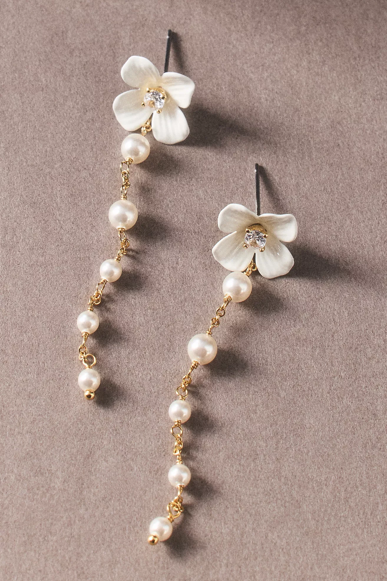 Close-up of a pair of earrings featuring white flower-shaped tops with rhinestones, dangling pearls, gold accents, and a chain design, displayed against a beige background.