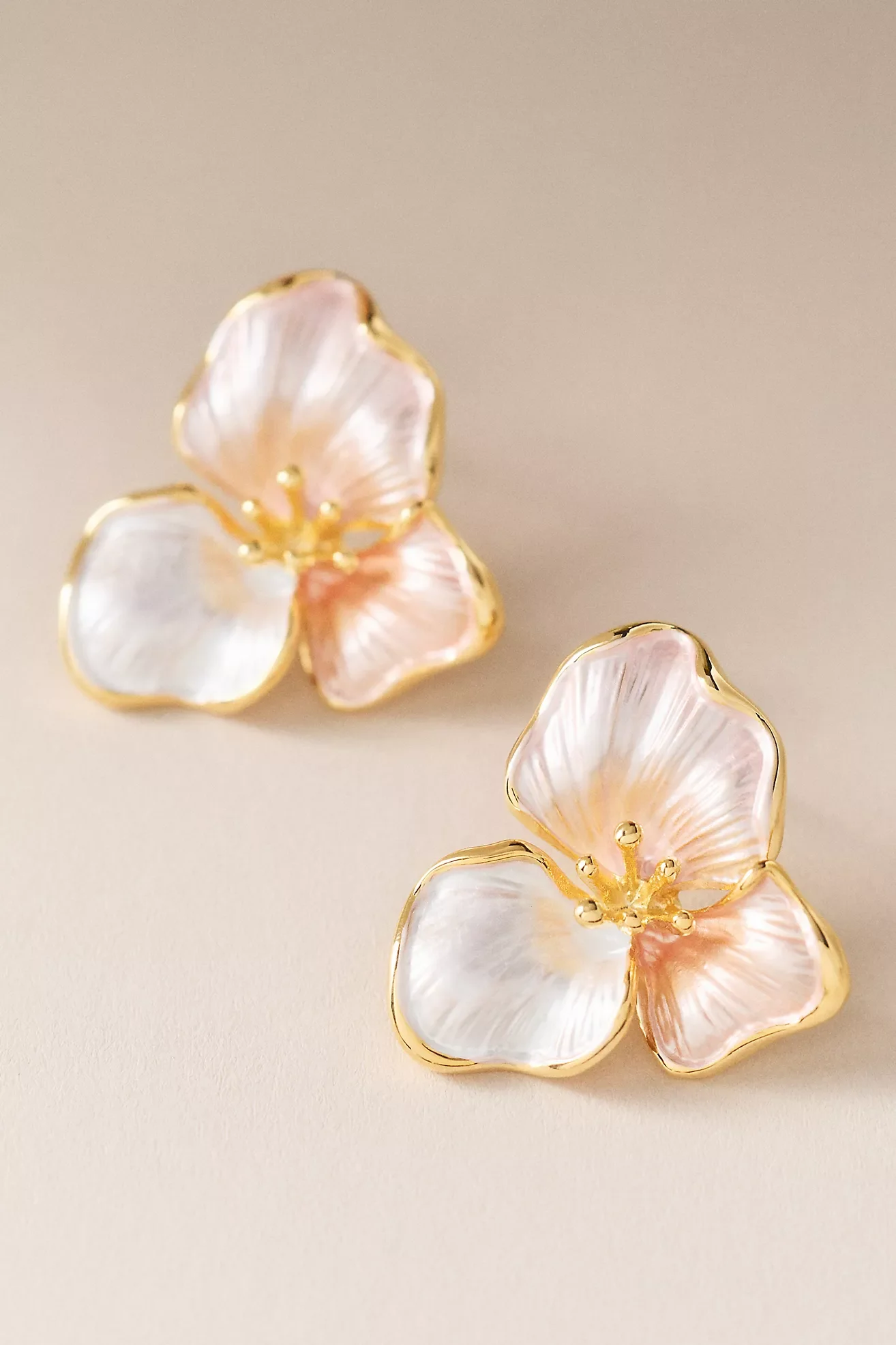 Close-up of three butterfly-shaped earrings with pink and white pearl-like petals and gold outlines on a beige background.