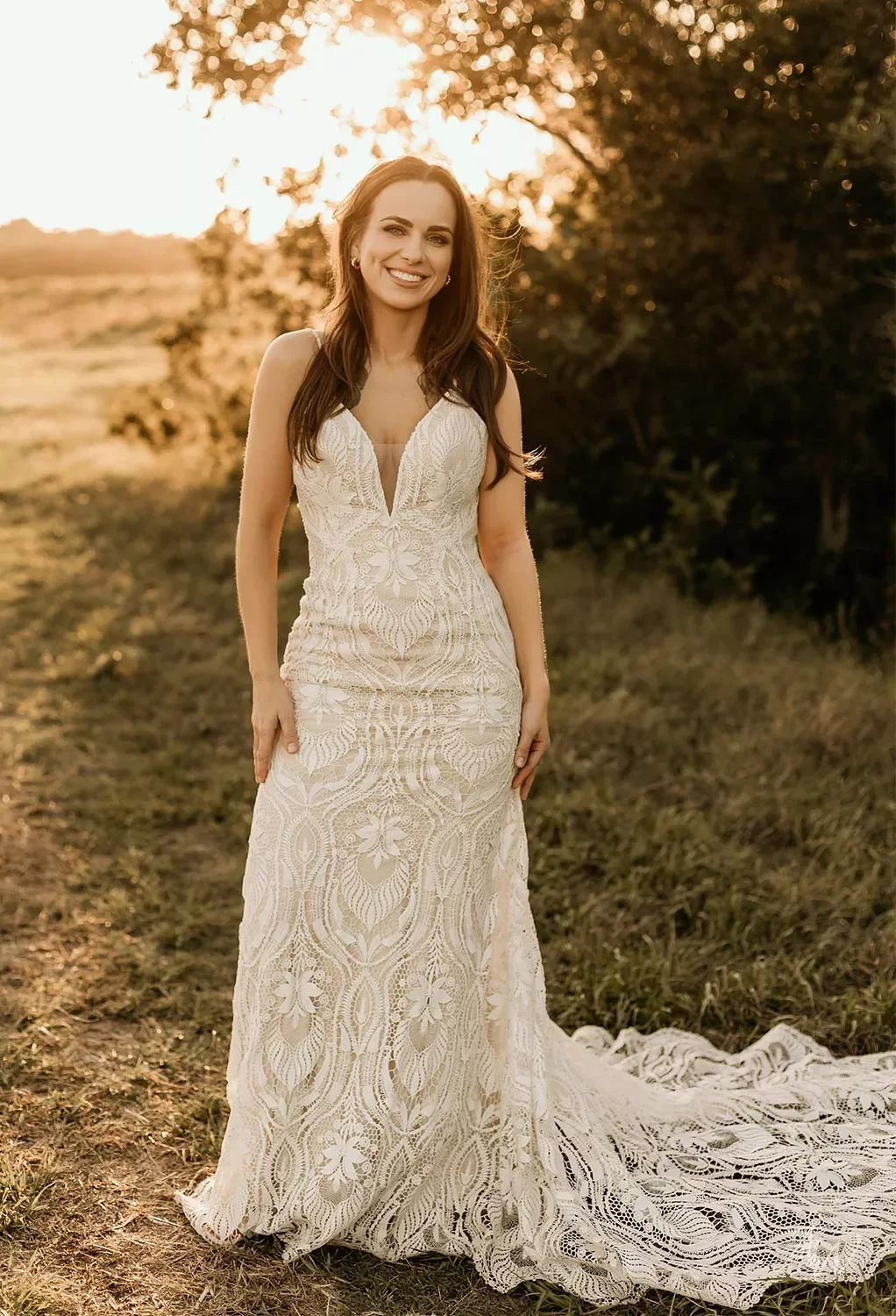 A woman in a white lace wedding dress standing outdoors on grass during sunset, smiling and facing camera.