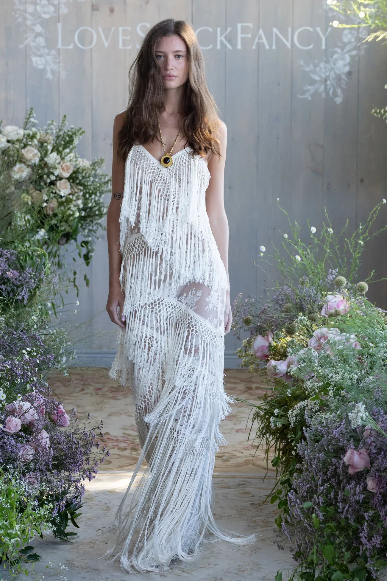 A woman walking on a runway surrounded by flowers, wearing a long, white, fringed dress and a gold necklace with a dark pendant.