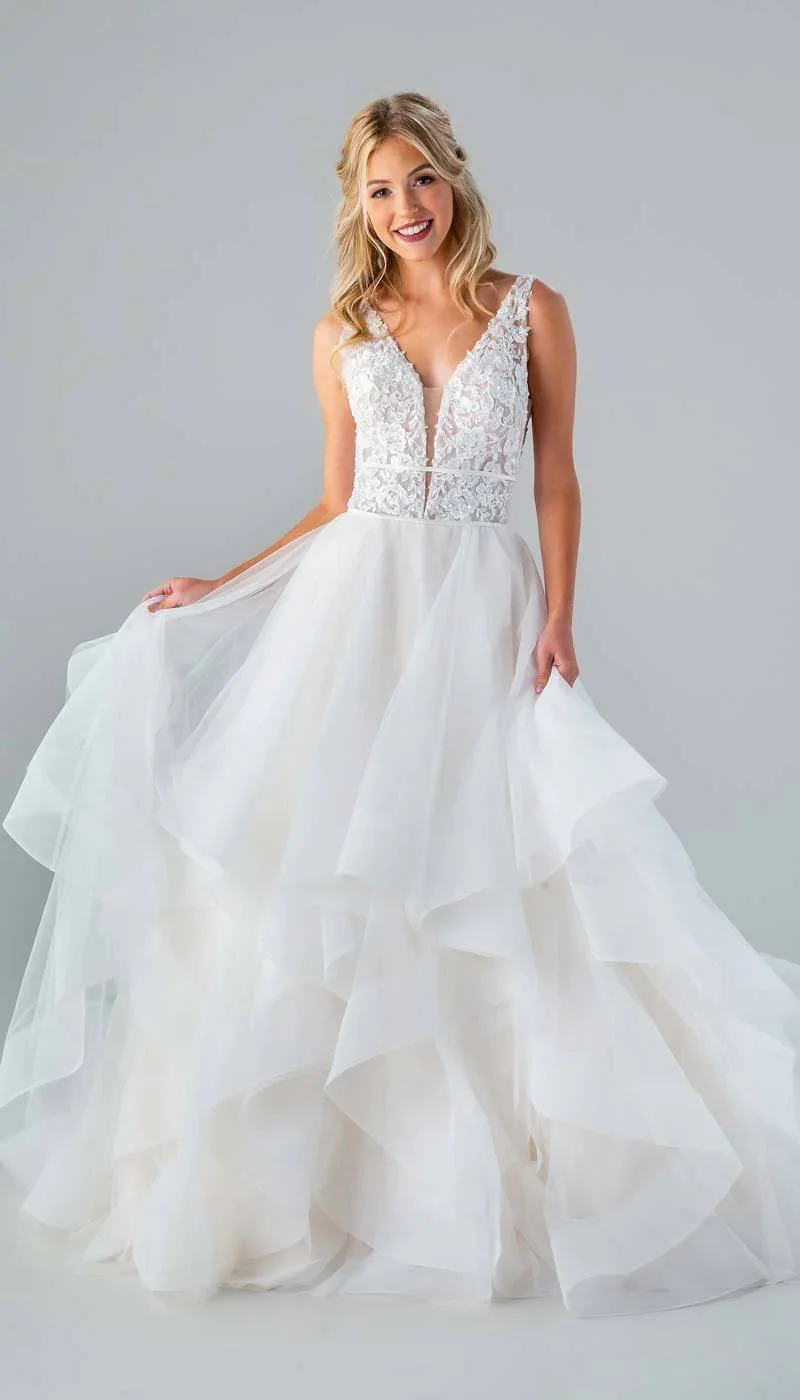 A smiling woman in a white wedding dress with lace bodice and layered tulle skirt posing against a plain grey background.