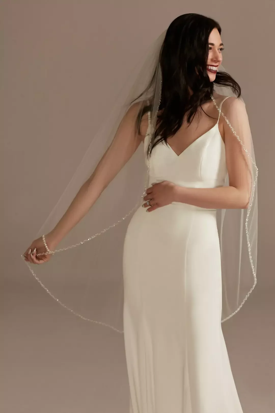 Woman in a white dress, wearing a veil, smiling, standing against a neutral background.