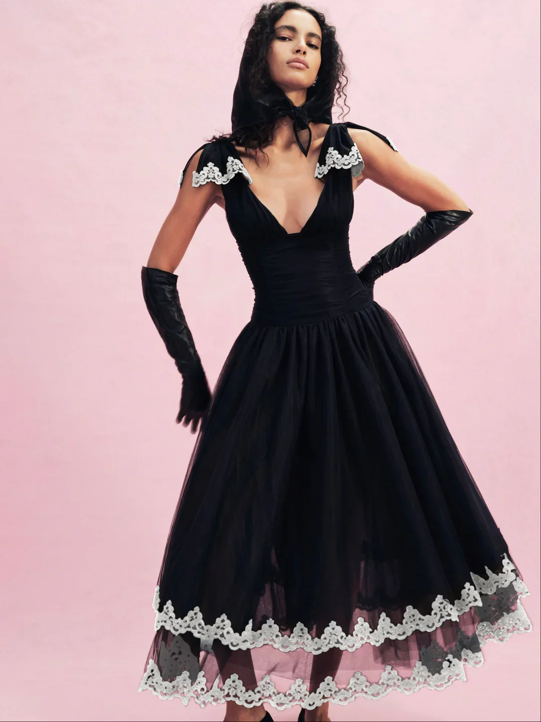 A woman in a black dress with white lace details and long black gloves, standing against a pink background.
