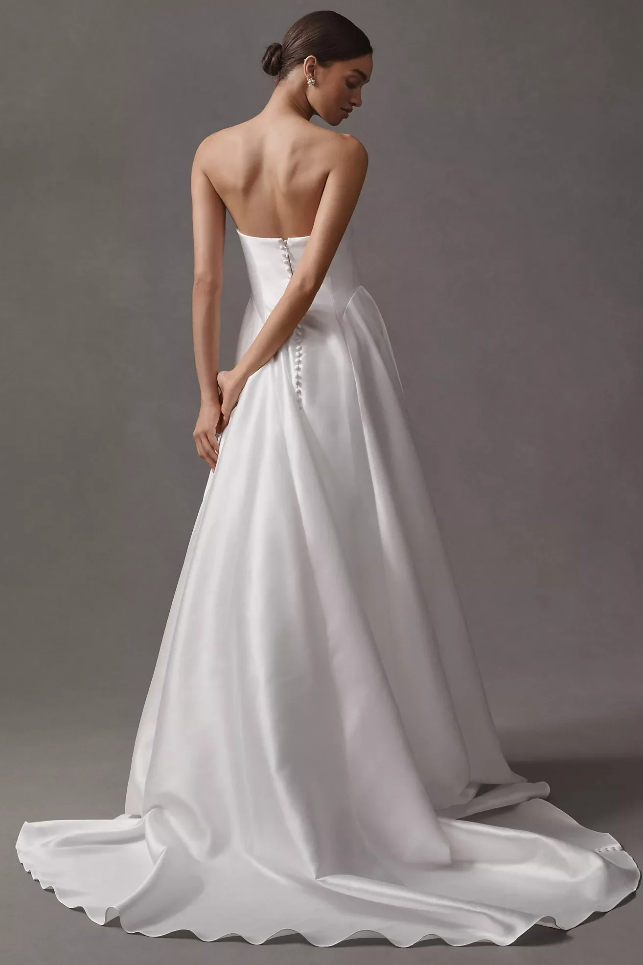 A woman in a white wedding dress standing against a plain gray background, with her back turned and looking to the side.