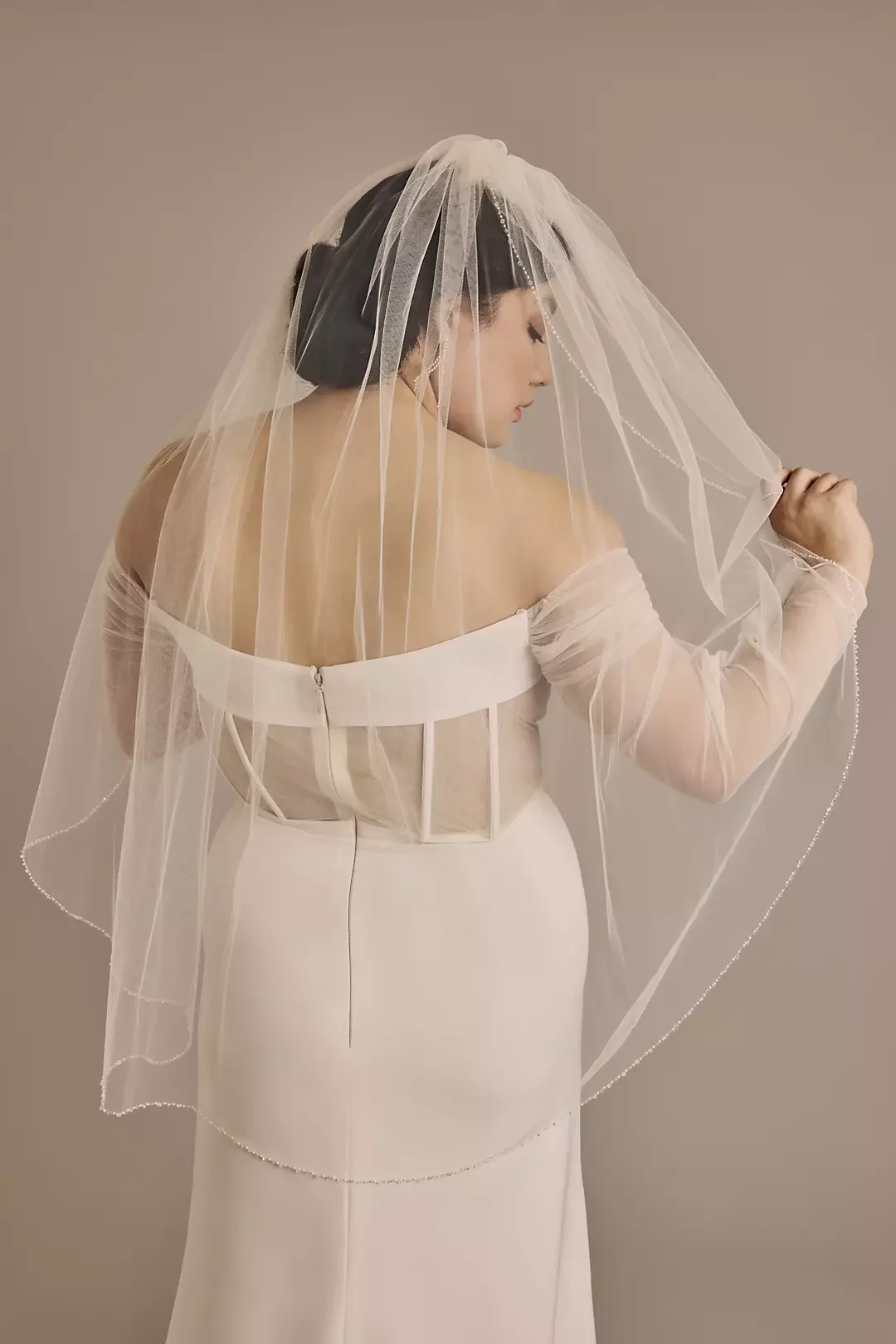 Woman in off-shoulder wedding dress with veil, turned to side, holding veil with one hand against neutral background.