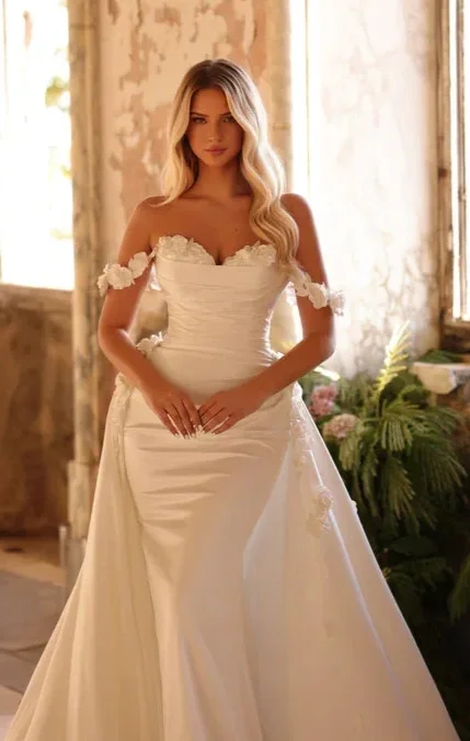 A woman in a white wedding dress stands indoors, with rustic walls and greenery in the background.