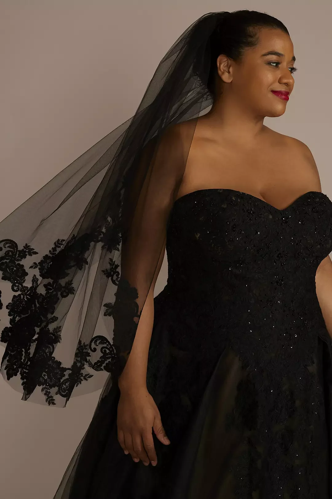 A woman in a strapless black gown with intricate embroidery, wearing a black veil with floral embroidery, standing against a neutral background.