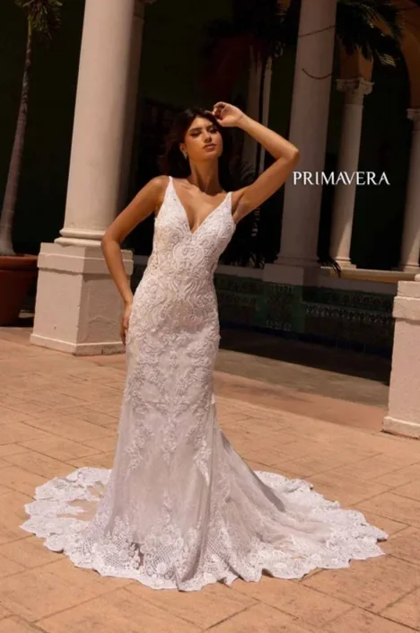 A woman wearing a white lace wedding dress with a train stands outdoors near white columns, with palm trees in the background, under a dark sky.