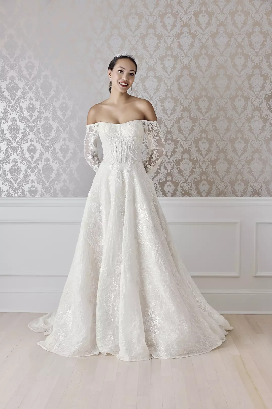 A woman in a white wedding dress standing indoors against a patterned wallpaper background.