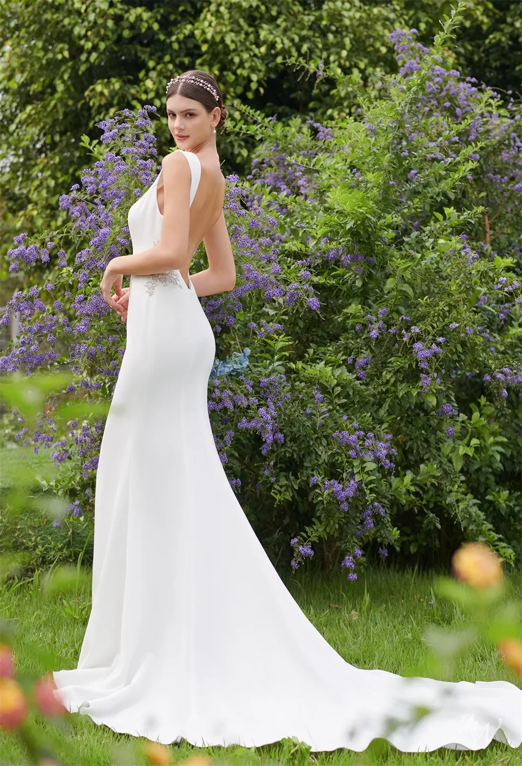 A woman in a white wedding gown standing outdoors in front of purple flowering bushes.