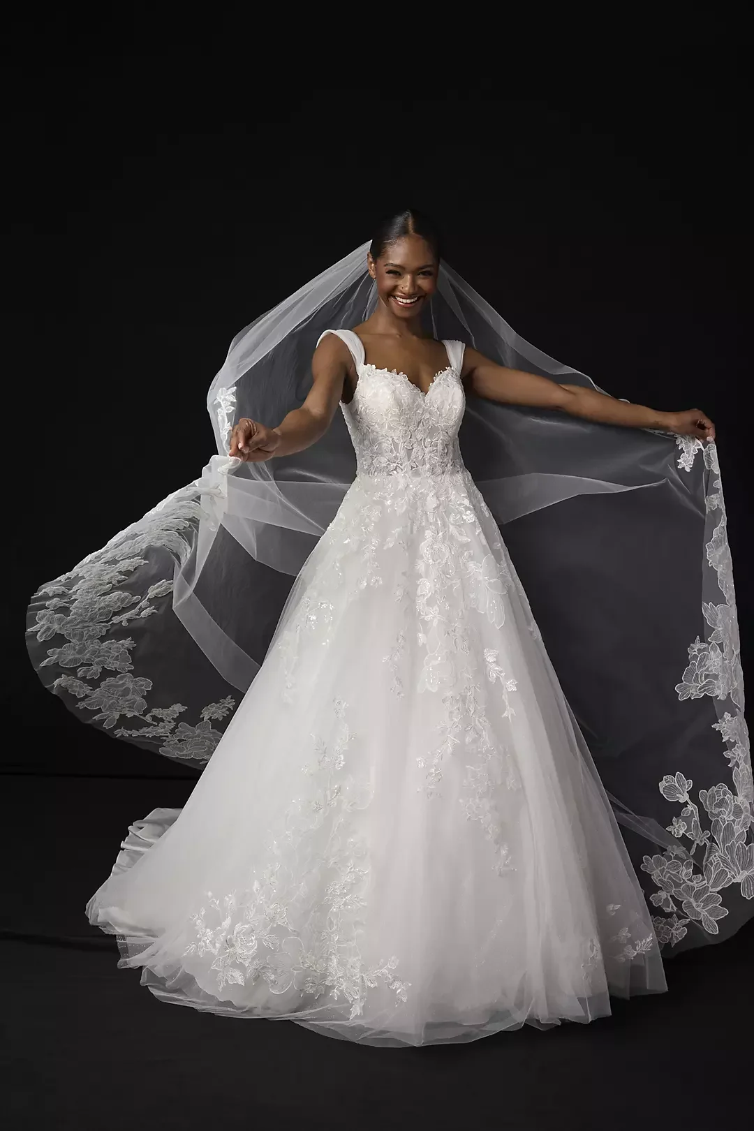 A woman wearing a white wedding gown with lace detailing, holding a sheer veil with lace edges, standing against a black background and smiling.
