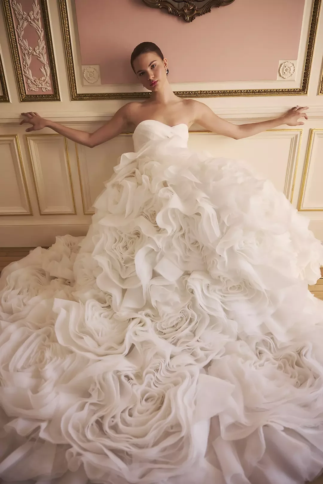 A woman wearing a strapless white wedding gown with a ruffled, voluminous skirt standing against a decorative wall.