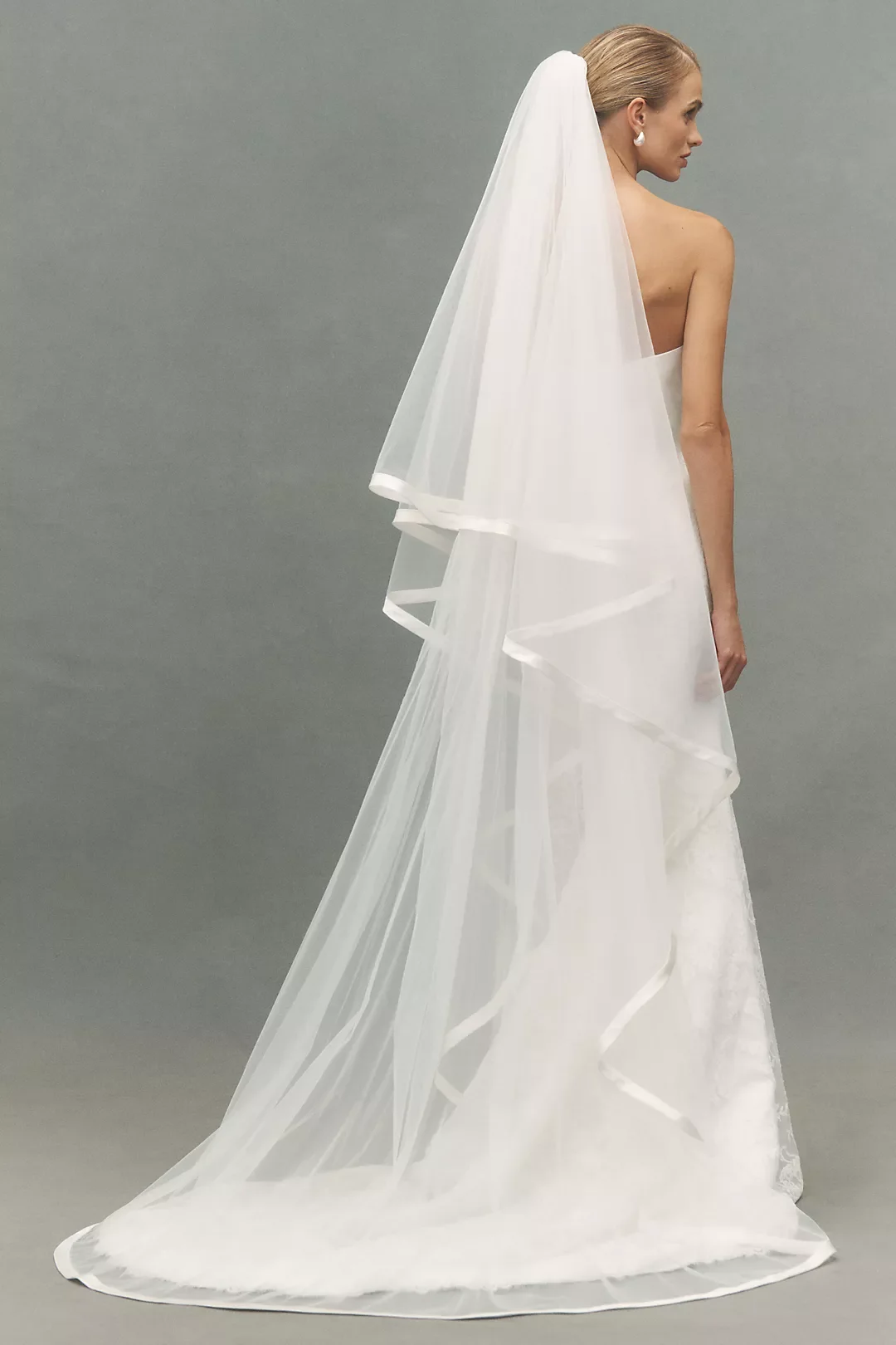 A woman in a white wedding dress with a long veil standing against a plain gray background, facing sideways.