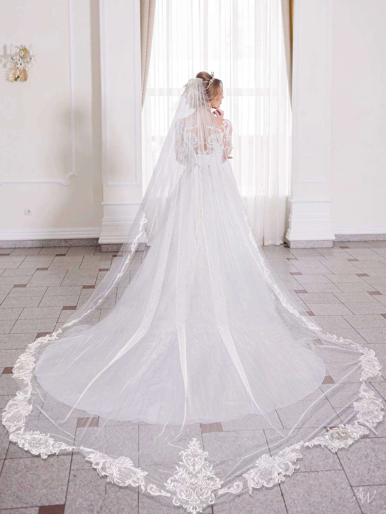 Bride in a white wedding gown with a long, embroidered cathedral veil standing in front of a window in a bright room.