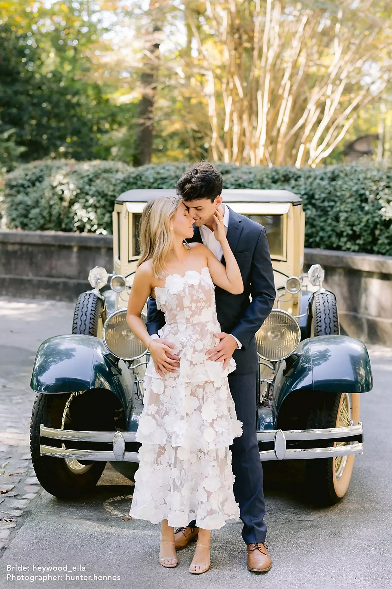 A bride and groom embrace in front of a vintage car outdoors with greenery and trees in the background, both smiling.