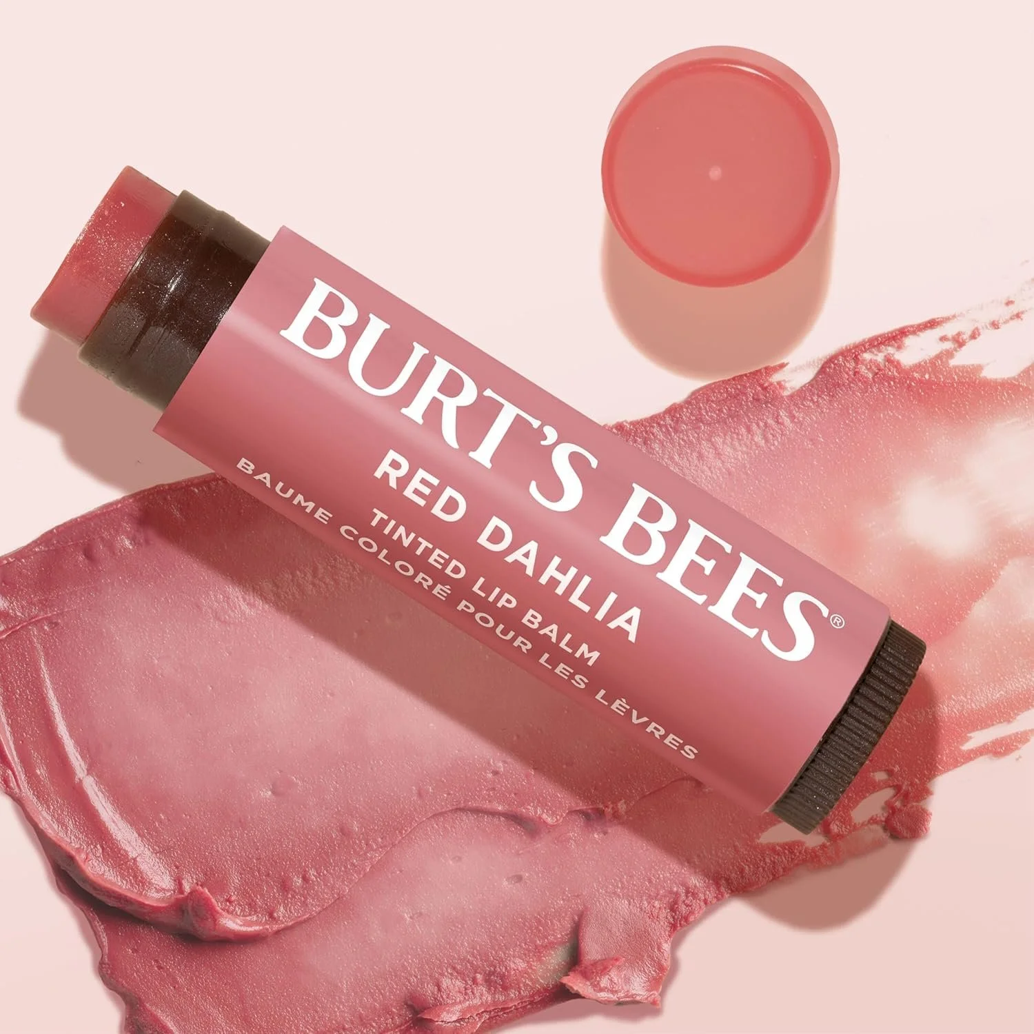 A Burt's Bees lip balm tube labeled 'Red Dahlia' resting on pink lipstick swatch with a small open cap beside it against a light pink background.
