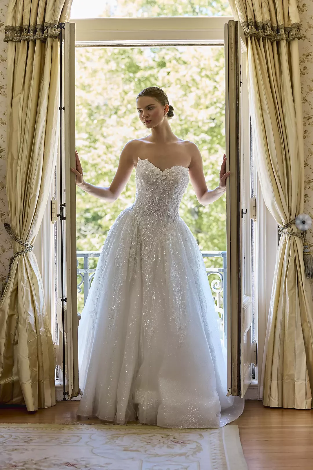 A woman in a strapless white wedding gown standing in a doorway, holding open cream-colored curtains with a scenic view of green trees outside.