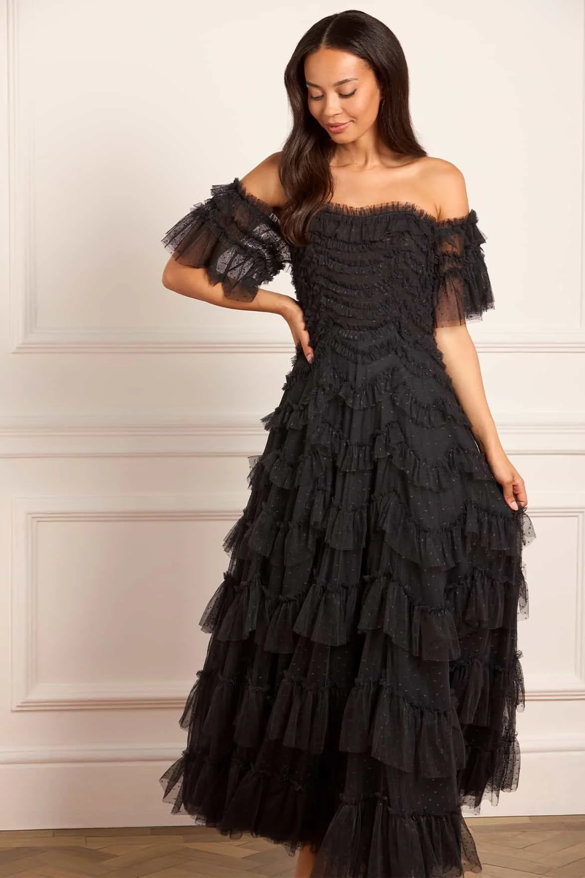 Woman in a black, off-shoulder, ruffled gown standing against a white wall with decorative molding.