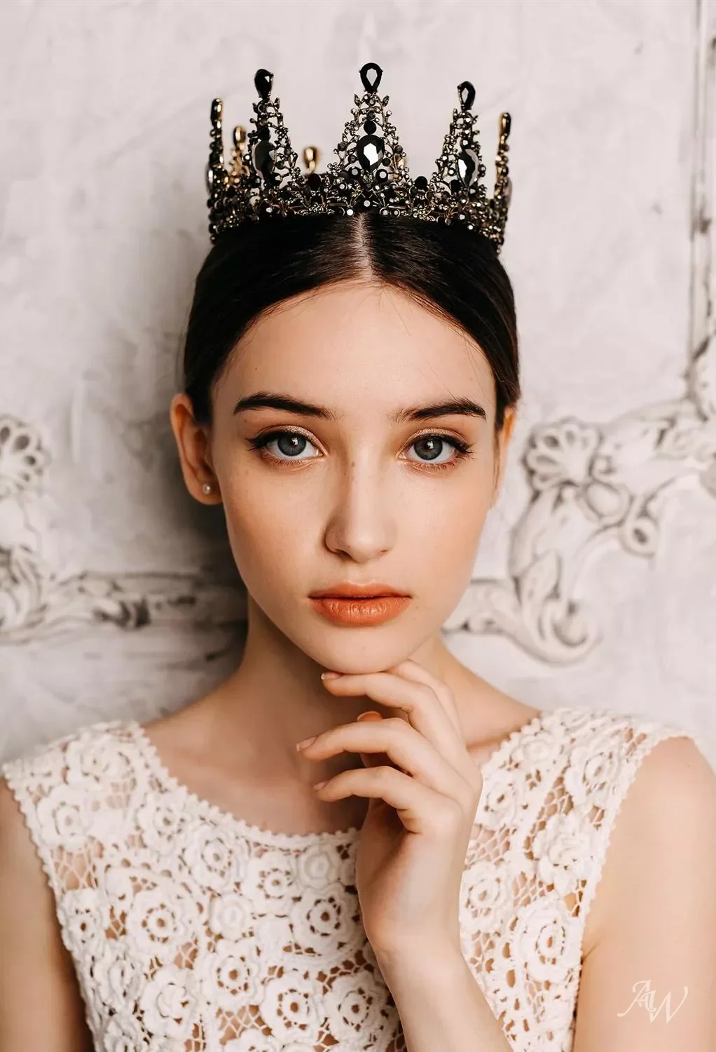 A young woman with blue eyes and dark hair, wearing a black ornate crown and a white lace sleeveless top, poses in front of a textured wall.