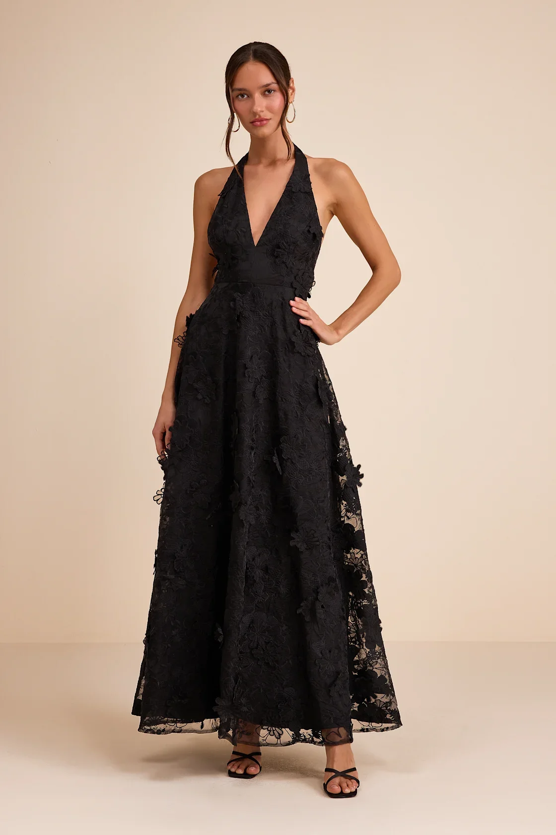 A woman in a long black lace dress with a deep V neckline, standing against a plain beige background.