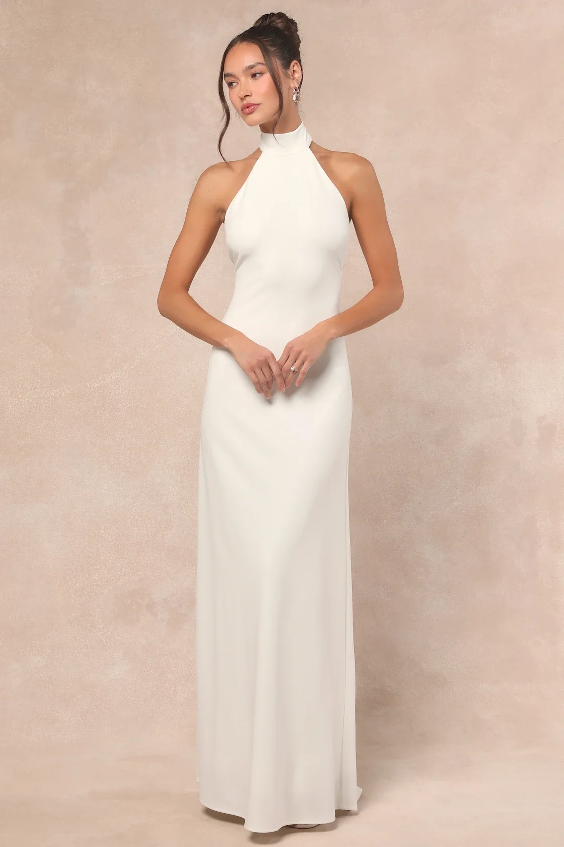 A woman wearing a white sleeveless, high-neck dress standing against a beige background.