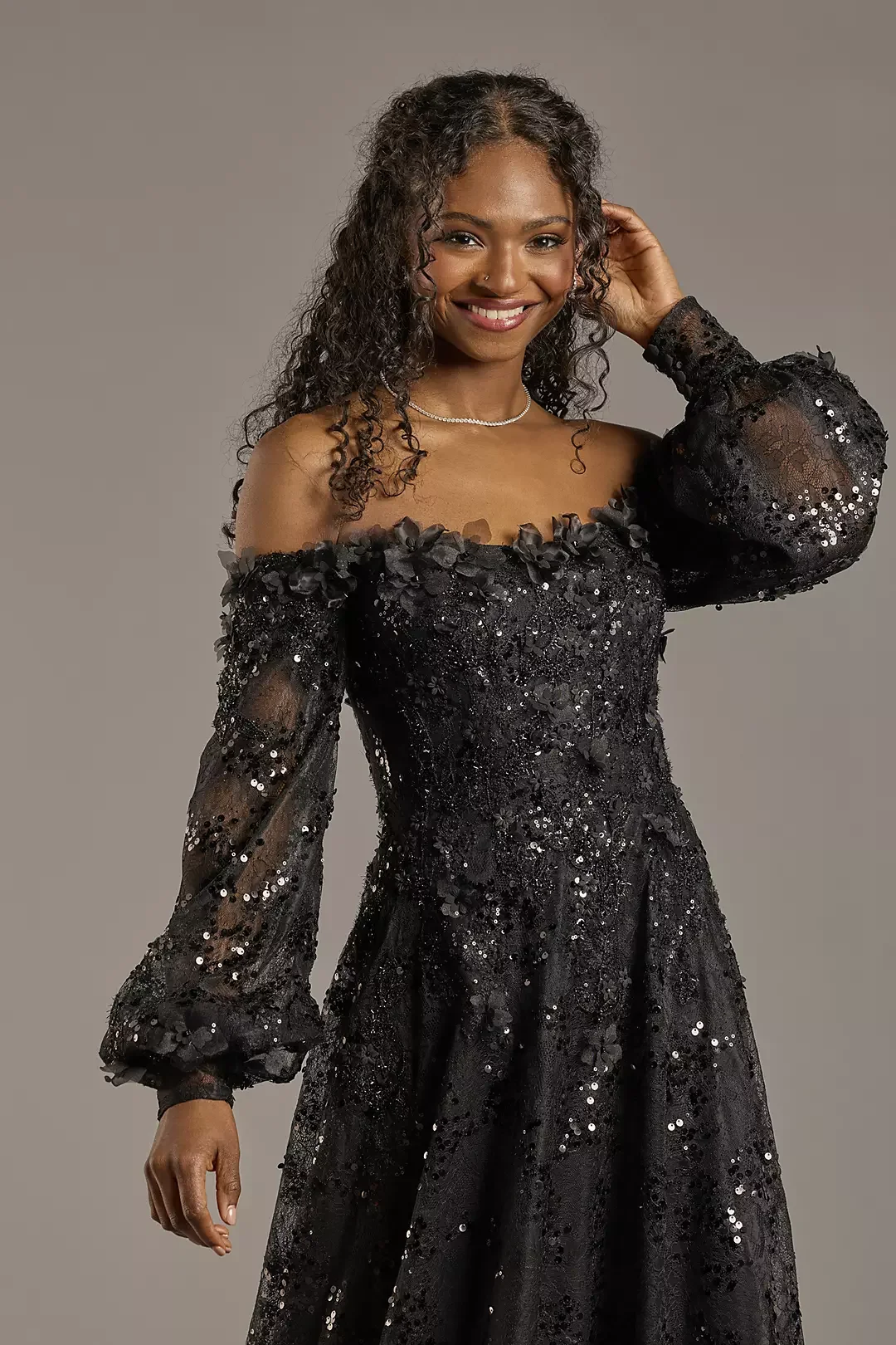 A woman with curly hair wearing a black sequined off-the-shoulder gown with sheer puffy long sleeves and a pearl necklace, smiling at the camera.
