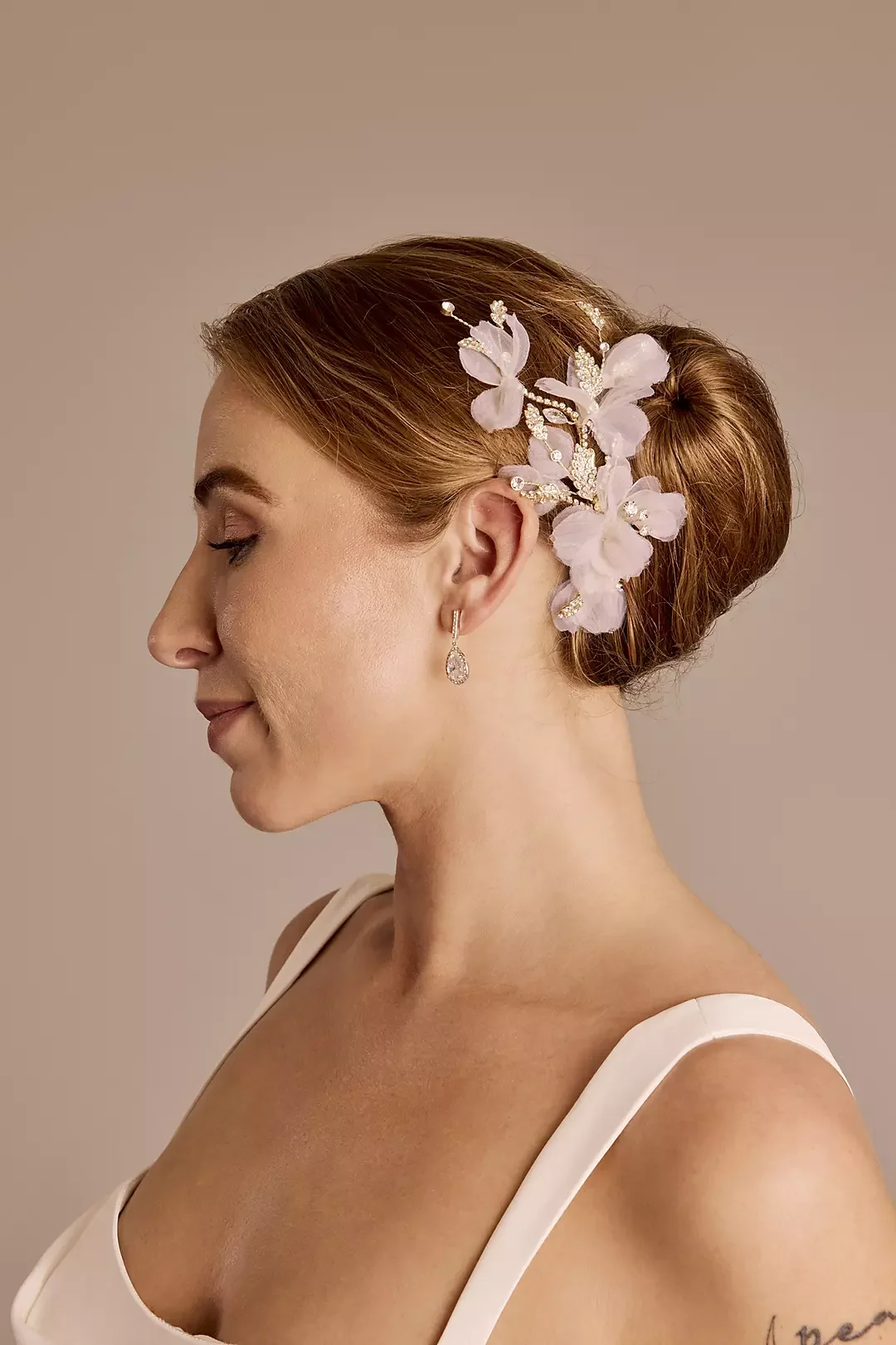 Side profile of a woman with red hair styled in a bun, wearing a floral hairpiece, a teardrop earring, and a white dress.