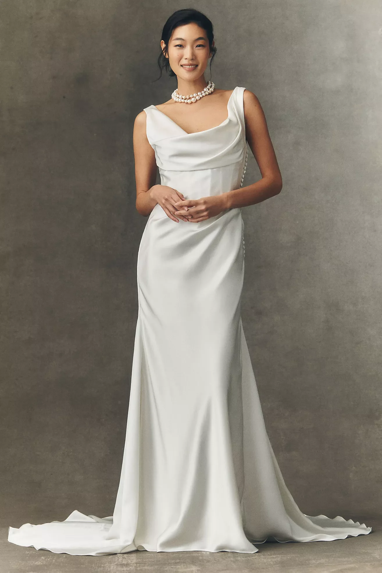 Young woman in a white wedding dress with a draped bodice and pearl necklace standing against a neutral background.