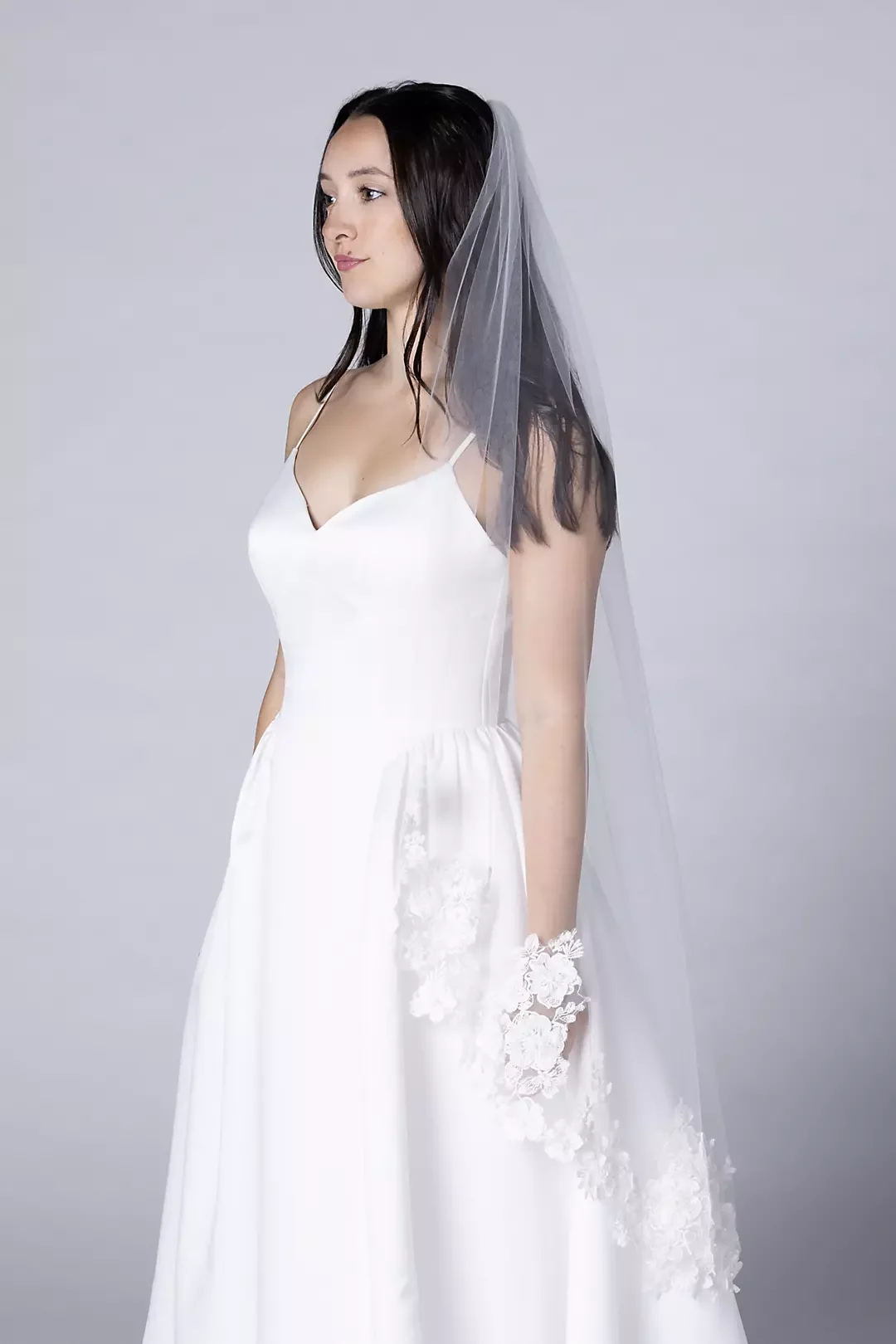 A woman in a wedding dress with a veil, standing sideways against a plain gray background.