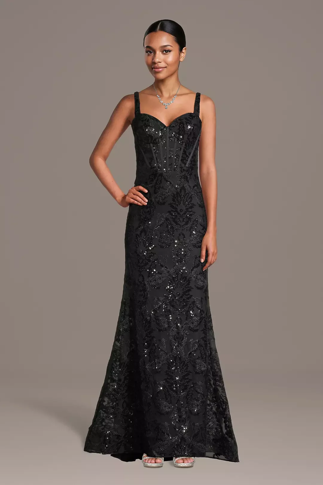 A woman in a long black sequined evening gown with intricate floral patterns, accessorized with a silver necklace and silver high-heeled sandals, standing against a plain background.
