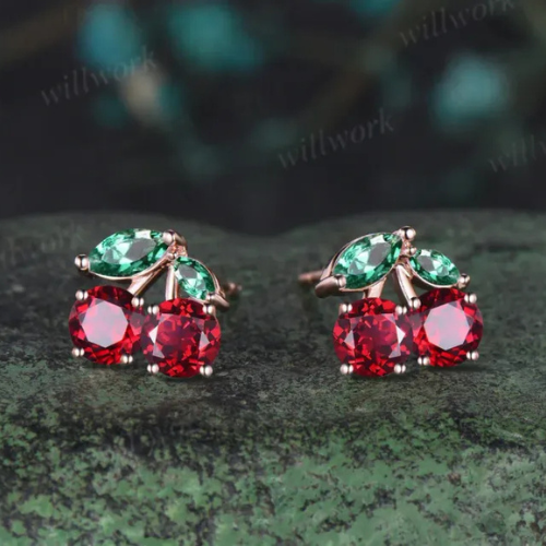 Pair of earrings with red gemstones and green leaf-shaped accents on a dark green textured surface. jewelry, cute jewelry, everyday jewelry, special occasion jewelry, ruby, diamond, emerald, gold