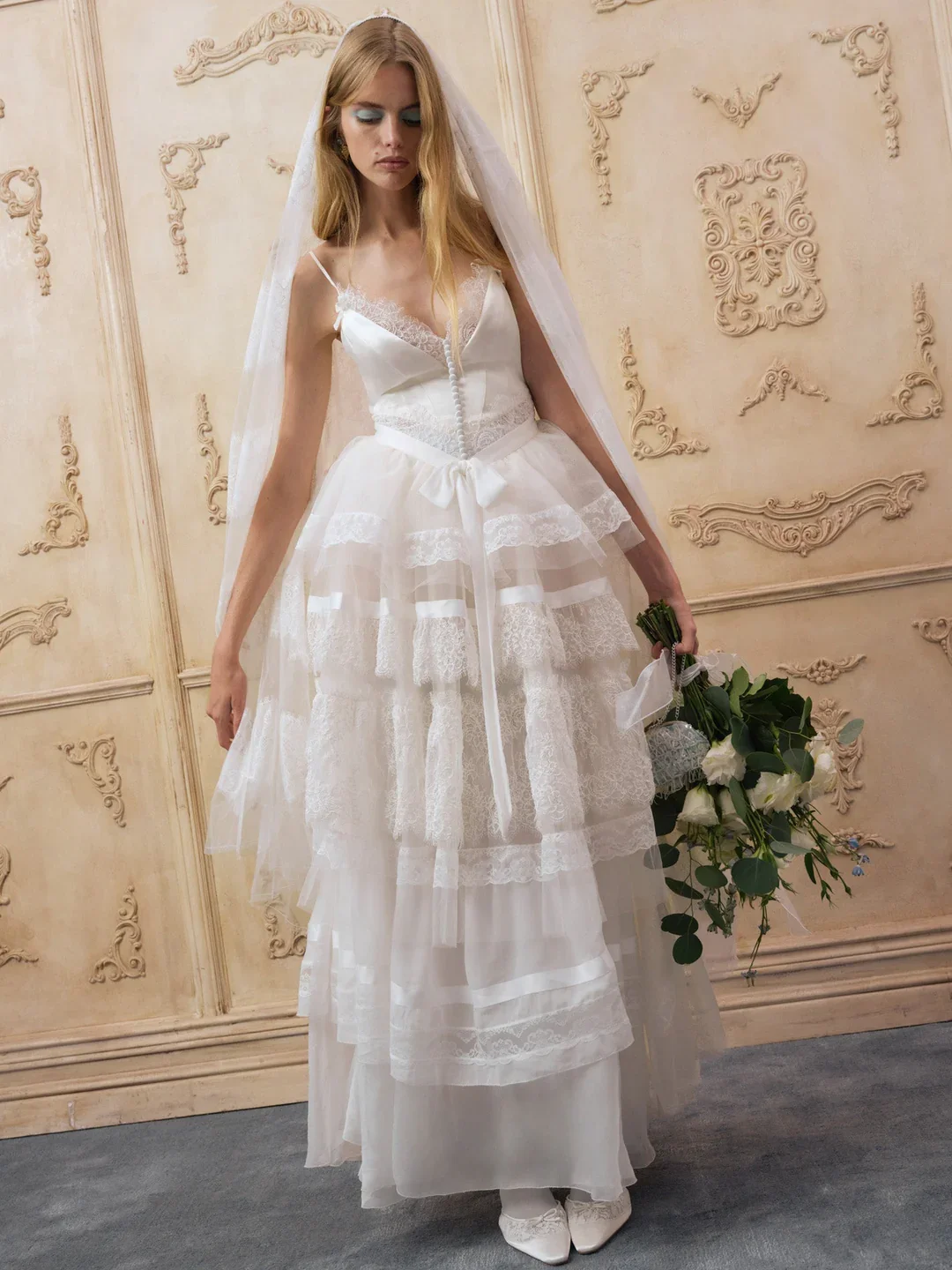 A woman in a white wedding dress with a lace and ribbon design, wearing a veil, holding a bouquet of flowers, standing against an ornate cream-colored wall.