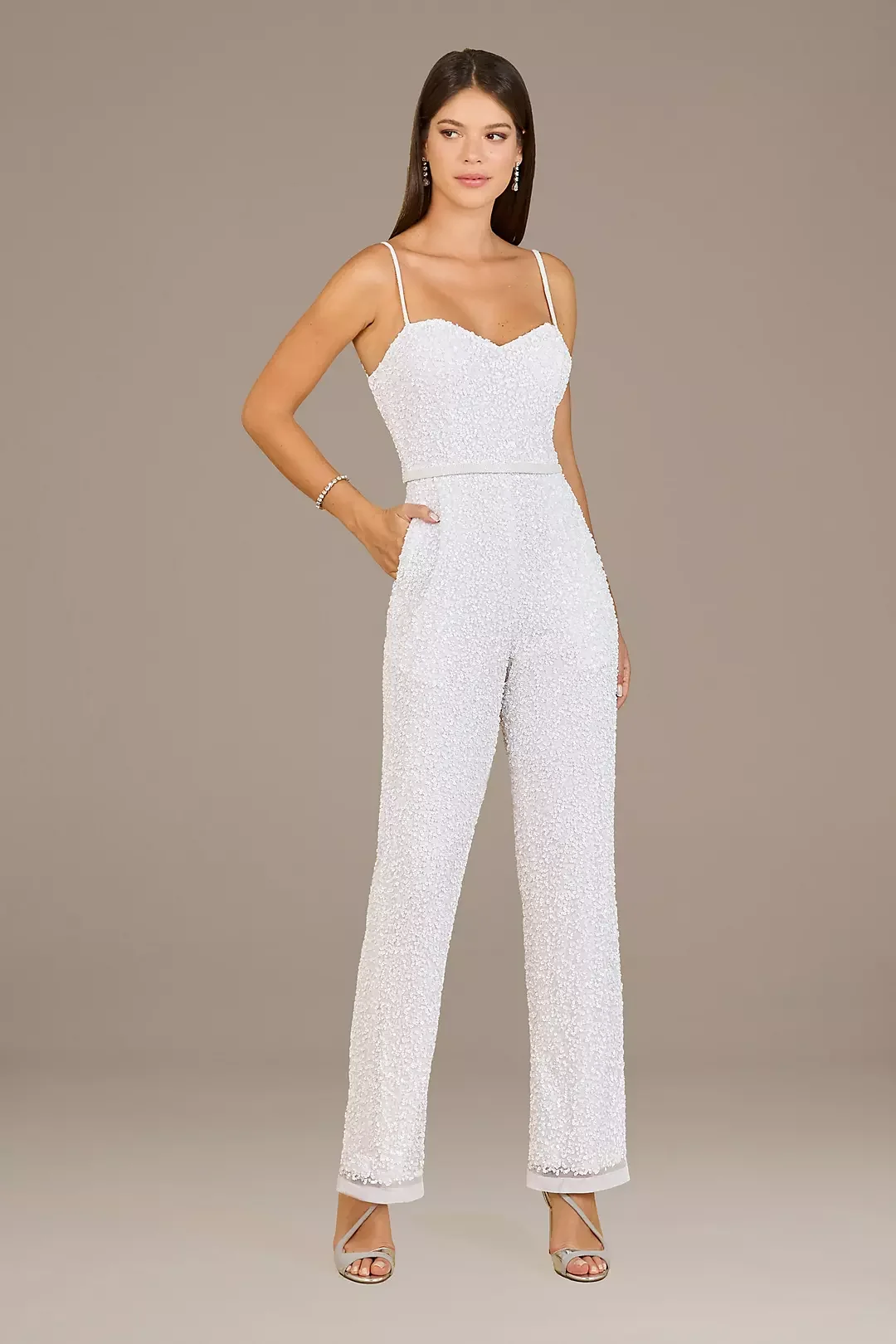 A woman wearing a white, textured jumpsuit with spaghetti straps and heels, standing against a plain background.