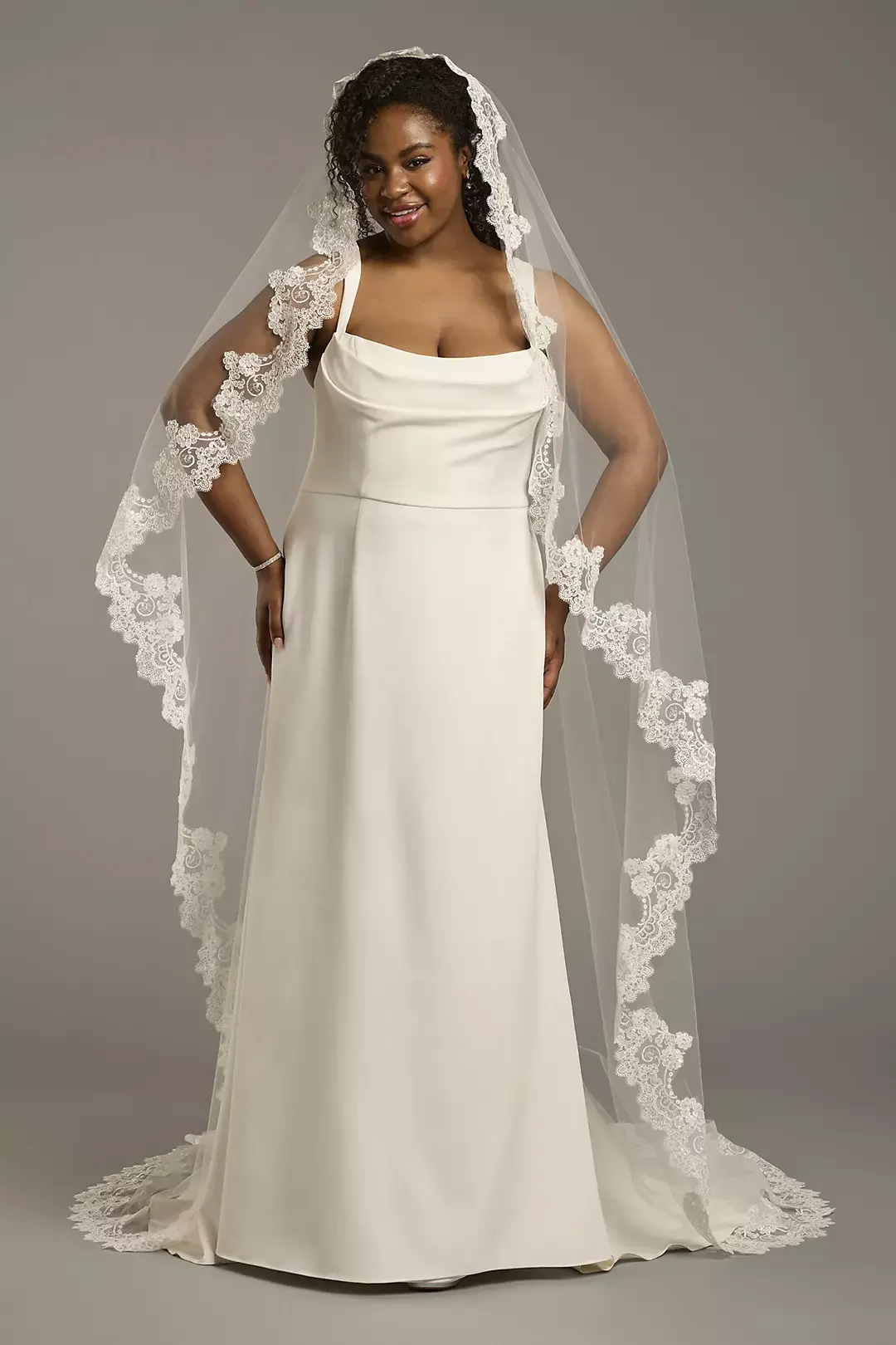 A smiling woman with dark curly hair wearing a white wedding dress and lace-edged veil standing against a gray background.