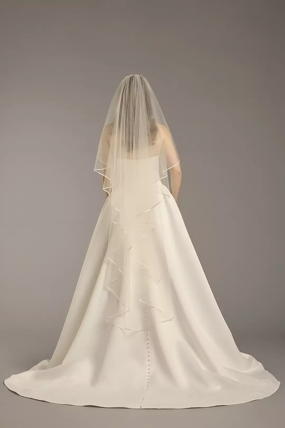 Back view of a bride in a white wedding gown and veil against a plain gray background.