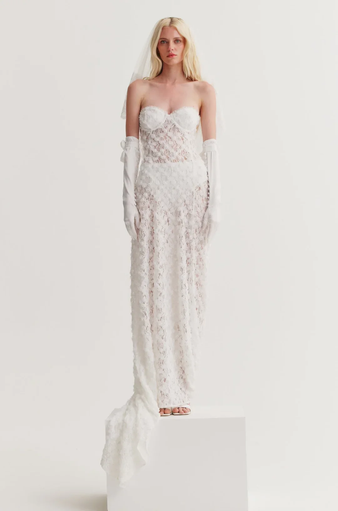 A woman in a long white lace wedding dress, wearing long white gloves, standing on a white pedestal against a plain white background.