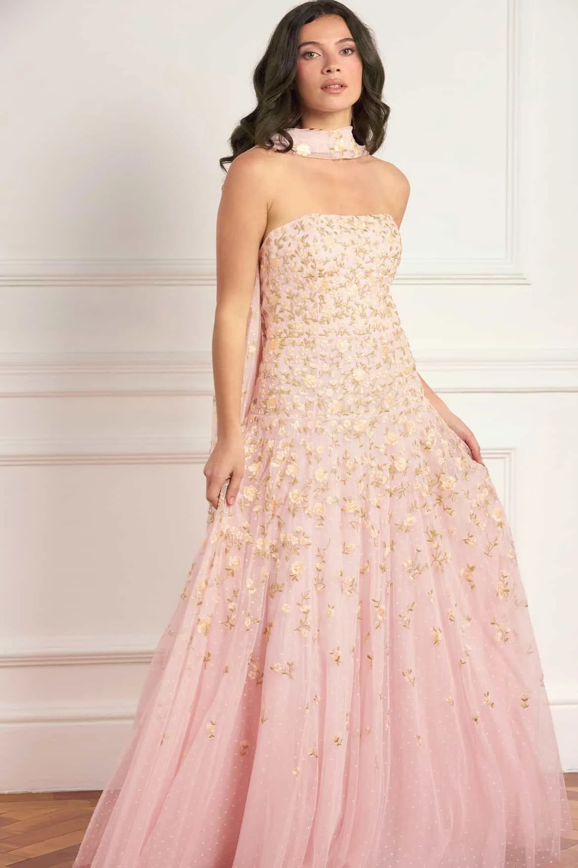 A woman with dark wavy hair wearing a pink gown with gold embroidery, standing in front of a white wall with decorative molding.
