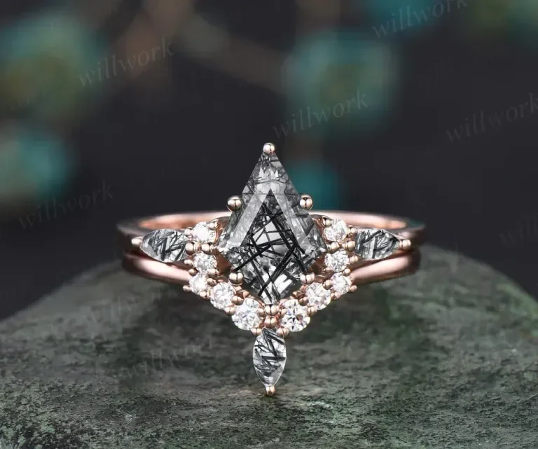Close-up of a rose gold ring with a large teardrop-shaped black and white diamond and small white diamonds surrounding it, set on a textured gray surface.